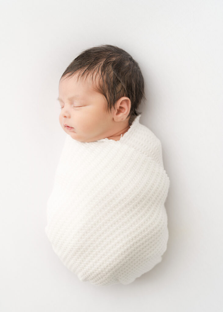 newborn baby boy with a headful of dark hair photographed at a newborn session on white
